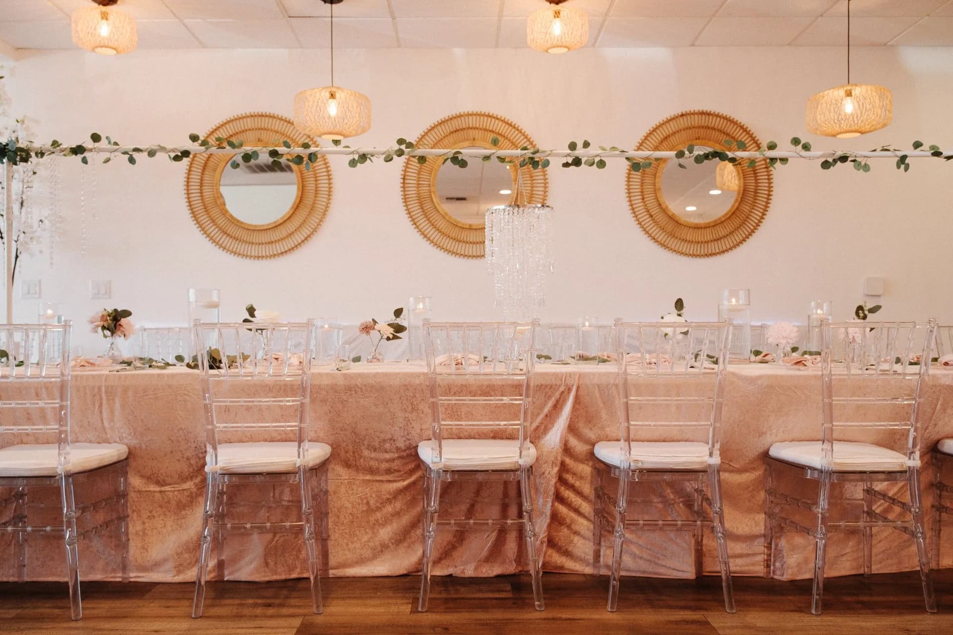 Front view of a blush head table with clear chairs, greenery, mirrors, and pendant lights