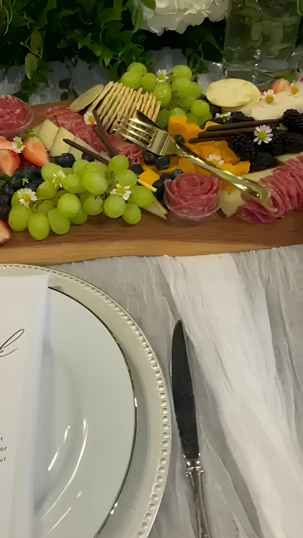 Individual charcuterie board with cheeses, fruit, meats, crackers, and white flowers beside a dinner plate