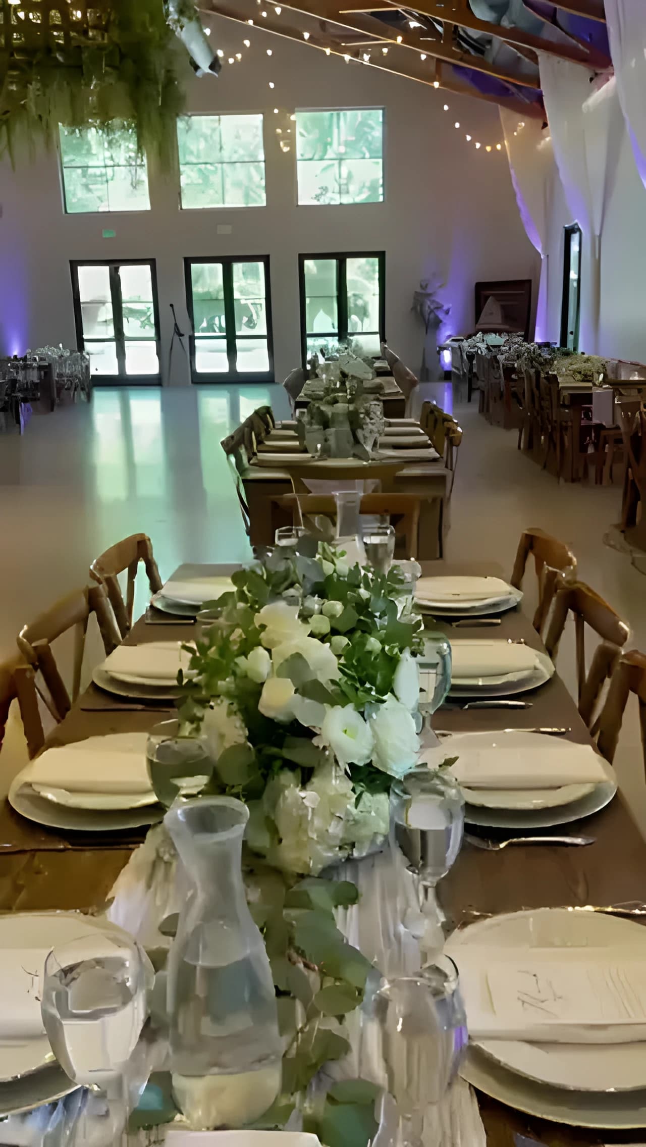 Long reception tables with white flowers, place settings, string lights, and blue uplighting