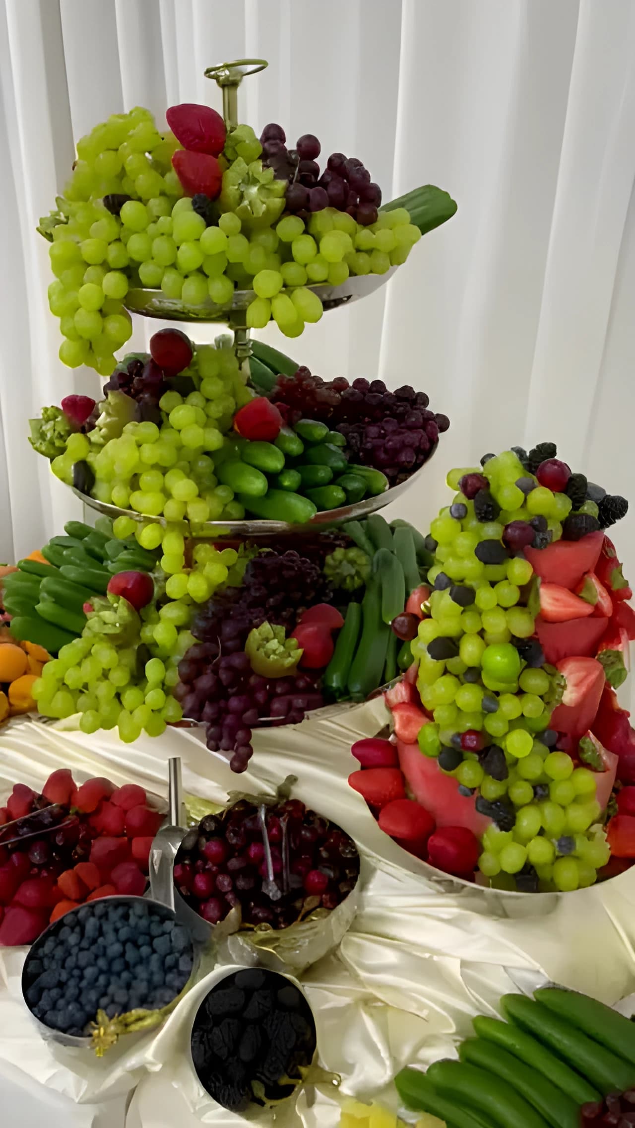 Tiered fruit display with green and red grapes, watermelon, berries, and pineapple