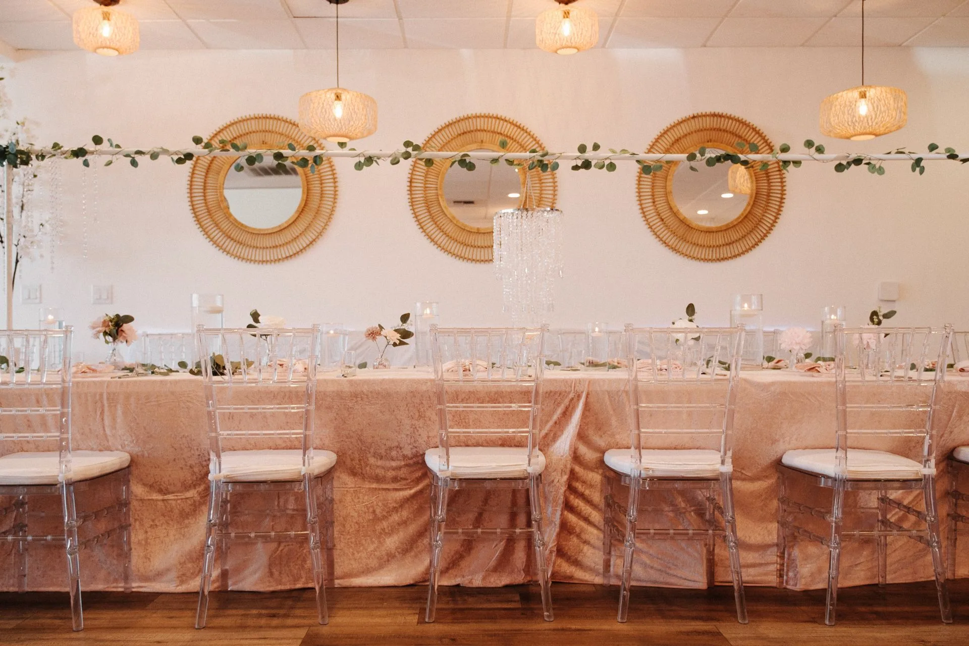 Front view of a blush head table with clear chairs, greenery, mirrors, and pendant lights