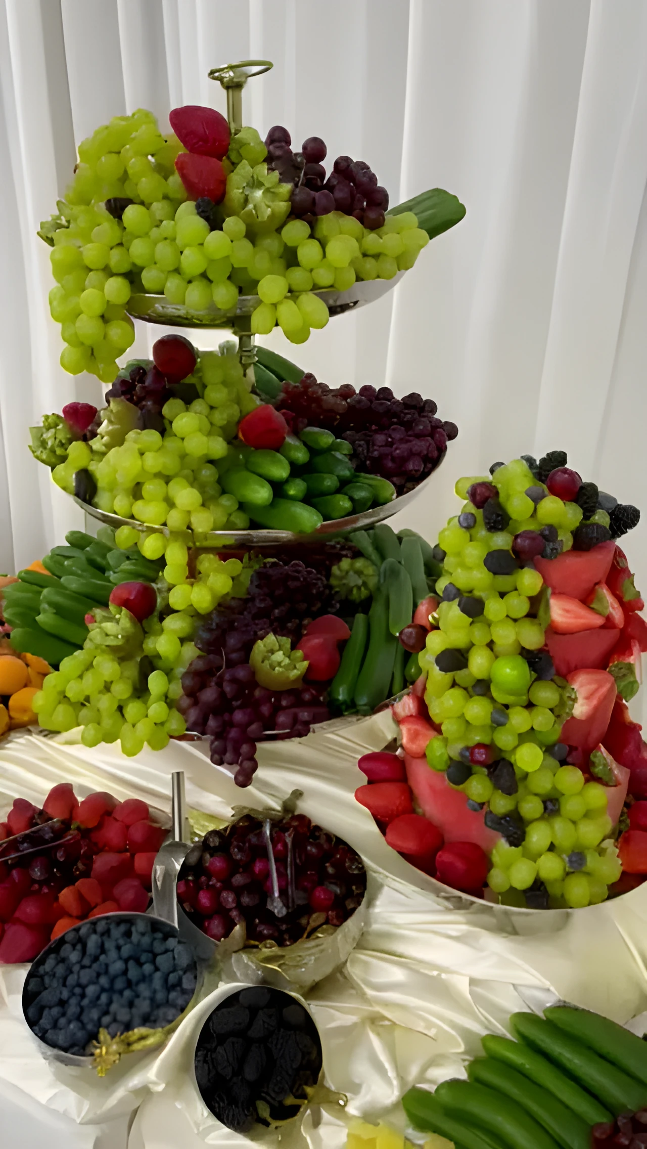 Tiered fruit display with green and red grapes, watermelon, berries, and pineapple